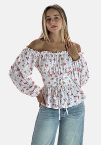 Weiße Bluse mit Blumenmuster, puffärmelig, gerafftem Ausschnitt und Schnürung in der Taille. Ausgestattet mit kleinen roten Blumen und einem ausgestellten Saum. Über Jeans getragen.