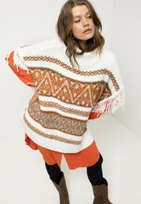 Weißer Strickpullover mit braunen geometrischen Mustern und Fransen, kombiniert mit einem orangefarbenen Kleid und schwarzen Leggings, getragen mit braunen Stiefeln.