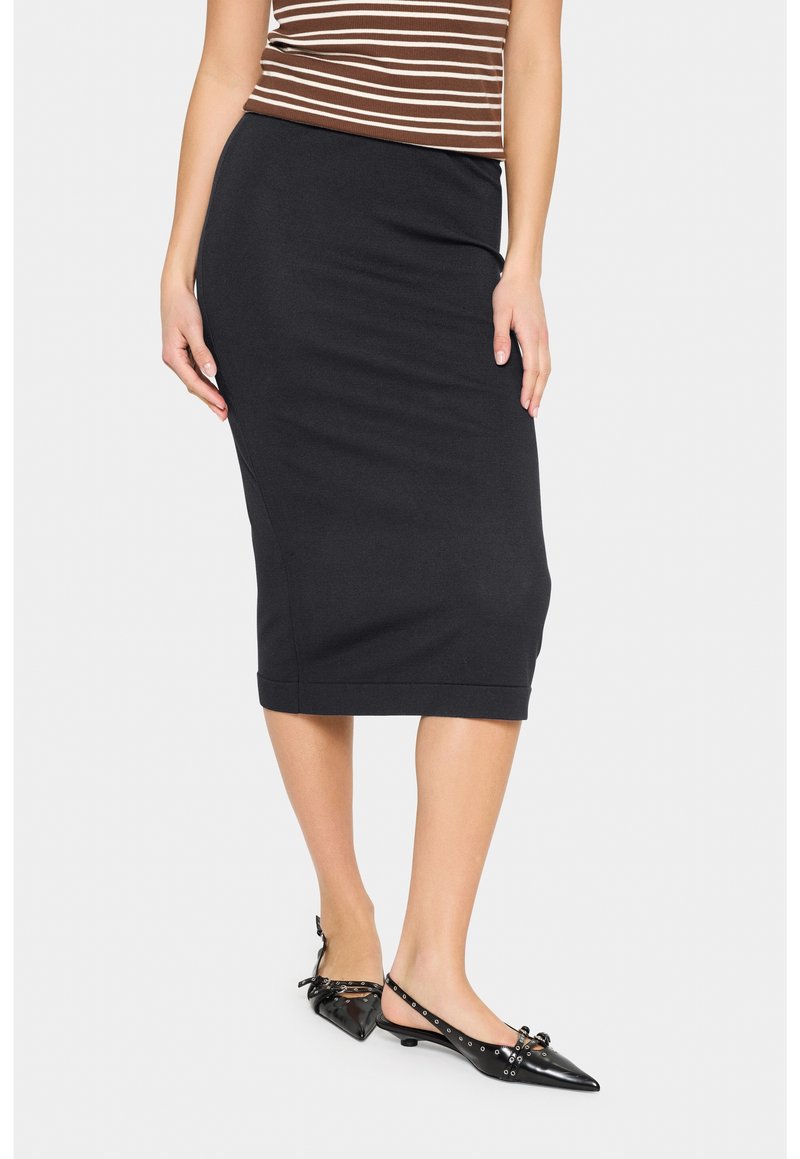 Femme portant une jupe crayon noire longueur genou, un haut rayé marron et blanc, et des ballerines slingback noires pointues avec des détails de nœud et œillets.