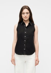 Γυναίκα με ίσια καστανά μαλλιά φοράει μαύρο αμάνικο πουκάμισο με κουμπιά και άσπρο παντελόνι, στέκεται μπροστ ά σε απλό ανοιχτό γκρι φόντο.