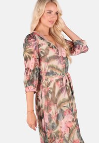 Blumengemustertes Wickelkleid aus durchscheinendem, leichtem Stoff. V-Ausschnitt mit Spitzeneinsatz, Dreiviertelärmel und Taillengürtel. Die Farben umfassen Pink und Grün.