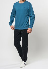 Modrý crewneck svetr vyrobený z měkkého materiálu, s malým logem na hrudi, v kombinaci s černými kalhotami a bílými teniskami.