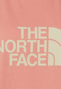 El logo de The North Face en beige sobre un fondo de tela rosa, mostrando texto parcial y el icónico símbolo del medio domo de la marca.
