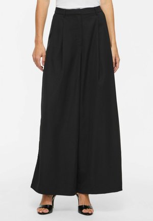 Femme portant un pantalon noir taille haute à jambes larges, associé à des escarpins noirs ouverts à l’avant, debout devant un fond blanc uni.