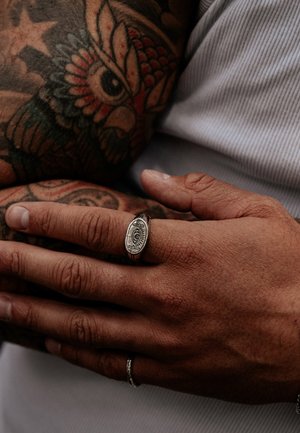 Silver signet ring with engraved design, worn on a finger of a hand with textured skin and visible tattoos. Natural lighting highlights details.