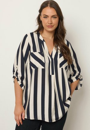 Mujer vistiendo una blusa suelta de rayas verticales negras y blancas, con las mangas arremangadas y dos bolsillos en el pecho, posando contra un fondo liso.