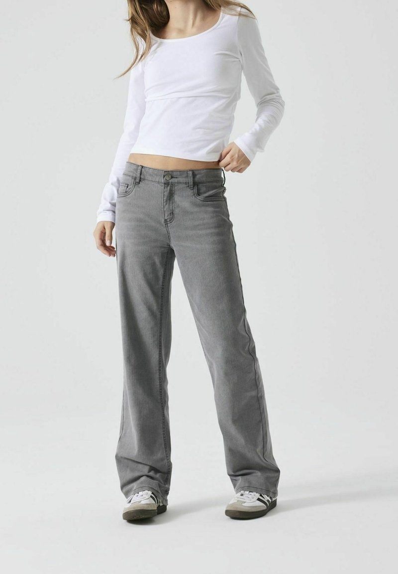 Femme portant un crop top blanc à manches longues, un jean large gris et des baskets blanches, se tenant contre un fond uni.