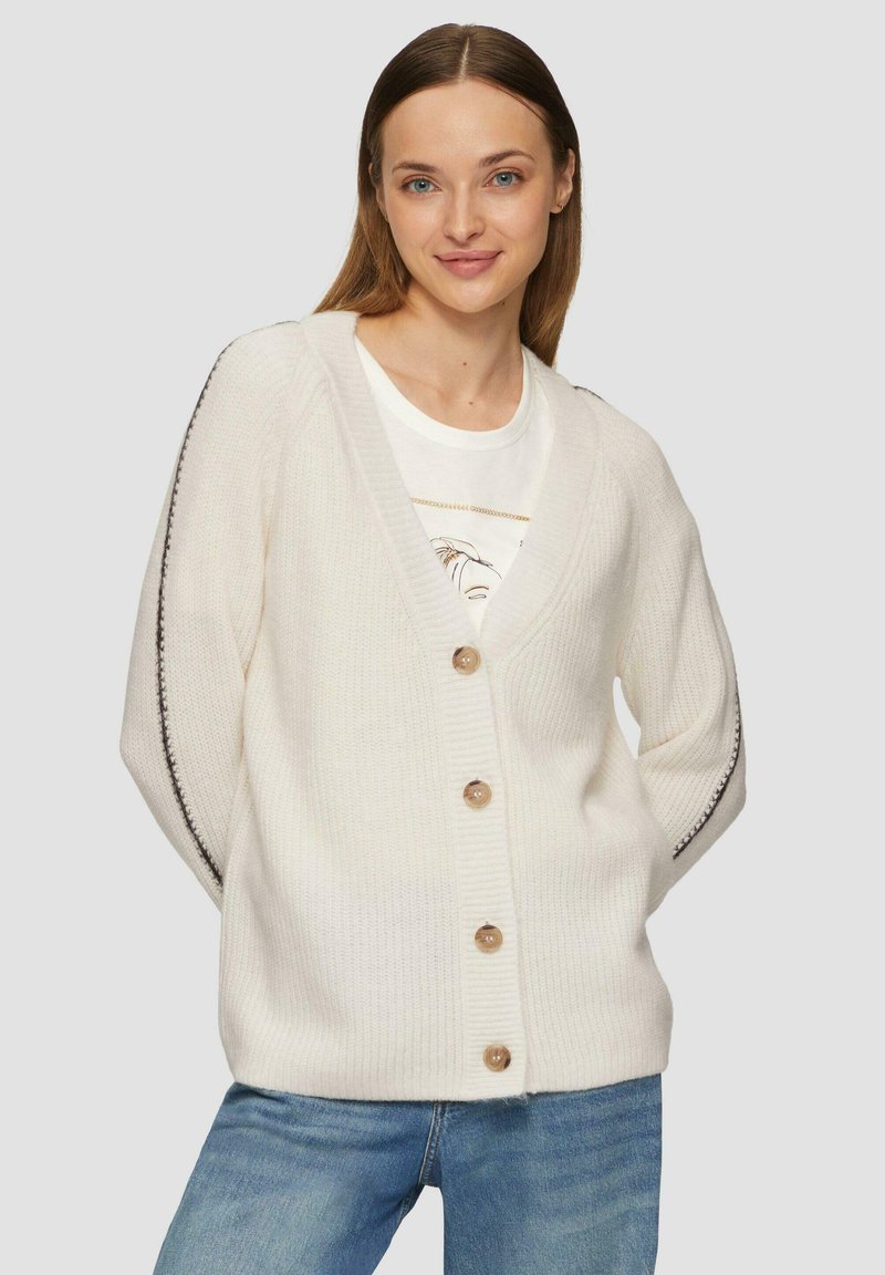 Młoda kobieta w białym prążkowanym kardiganie z guzikami i czarnym przeszyciem po bokach, ubrana w niebieskie dżinsy i białą koszulkę z nadrukiem pod spodem.