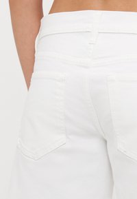 Pantalones cortos de mezclilla blanca con una cintura alta, que cuentan con dos bolsillos traseros y detalles de costura sutiles. La tela tiene una textura suave.