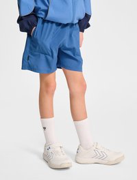 Blå idrottsshorts med en slät textur, utrustad med sidofickor och resårmidja. Kombinerat med vita sneakers och knähöga strumpor.
