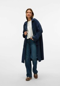 Marinblå regnjacka med huva och tryckknappar, lätt material, kombinerad med en vit skjorta, blå vida jeans och bruna skor.