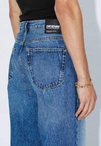 Blue denim jeans with a relaxed fit, featuring a back pocket and a black brand label. A gold bracelet is worn on the wrist.