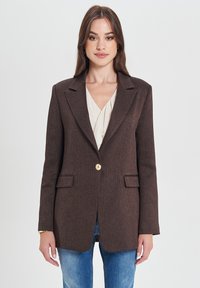 Blazer marrone a trama con un singolo bottone dorato, due tasche frontali e colletto a scollo, indossato sopra una blusa crema e jeans blu.