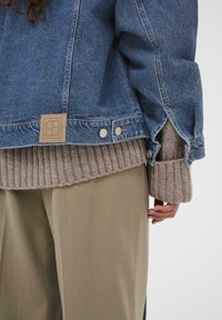 Traper jakna svijetloplave boje, s smešnim logotipom i metalnim dugmadi. Nosena preko rebrastog smešnog džempera i bež hlača.