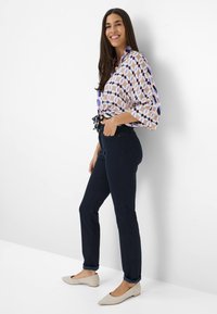 Geprinte blouse met geometrisch patroon in paarse en roze tinten, gecombineerd met marineblauwe slim-fit broek en beige puntige flats.