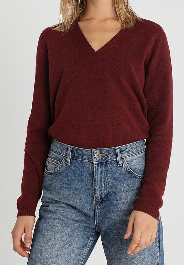 Femme portant un pull bordeaux ajusté à col en V, rentré dans un jean bleu taille haute, sur un fond uni.