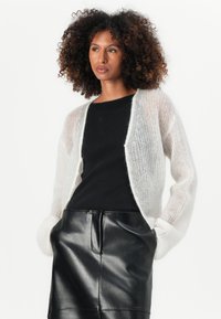 Hvit, gjennomsiktig, strikket cardigan med lange ermer og avslappet passform, brukt over en svart ribbet topp og et svart skinnskjørt.