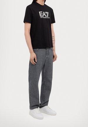 Homme portant un t-shirt noir EA7 Emporio Armani, un jean gris, des baskets blanches, se tenant avec les mains détendues le long du corps contre un fond uni.
