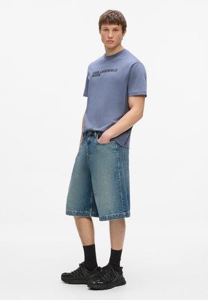 Młody mężczyzna w niebieskim t-shircie Karl Lagerfeld Jeans, krótkich dżinsowych spodenkach do kolan, czarnych skarpetkach i czarnych trampkach, stojący z rękami w kieszeniach.
