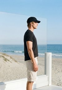 Jeune homme portant une casquette noire, un t-shirt noir et un short beige, debout face à l'océan sur une plage de sable derrière une rambarde en verre.