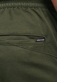 Pantalones cortos verde oliva con textura suave, que cuentan con un bolsillo lateral y un dobladillo cosido. Tiene una etiqueta negra de "VOLCOM" en el bolsillo.