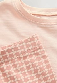 Lys pink T-shirt med en ribbet rund hals; har en lomme med et ternet mønster i mørkere pink og hvide syninger.