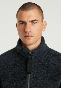 Giacca in pile di colore grigio scuro, con colletto alto, chiusura con zip e superficie texturizzata con una finitura morbida e pelosa.