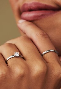 Twee ringen op een hand: een zilveren solitair met een heldere steen en een gestructureerde band met ronde accenten. Huidtint zichtbaar onder de ringen.