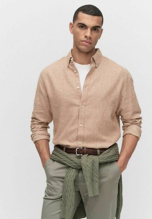 Chemise en lin marron clair avec un design à boutons, associée à un t-shirt blanc et un pull en laine vert noué autour de la taille. Pantalons beiges et ceinture marron.