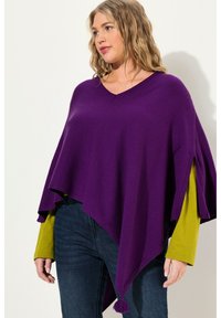 Poncho viola con design a V, realizzato in tessuto morbido. Presenta una vestibilità ampia, fessure laterali e un'applicazione di frange all'orlo. Abbinato a una maglietta verde.