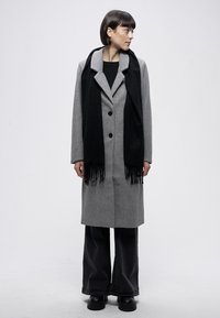 Grauer Wollmantel mit schwarzem Schal, ausgestattet mit Schalkragen, Knopfverschluss und Fransen-Details. Das Modell trägt weite schwarze Hosen und Stiefel.