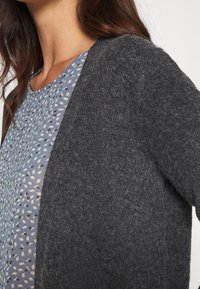 Femme portant un cardigan gris pelucheux par-dessus un haut bleu avec un petit motif blanc et foncé, montrant le haut du torse et le cou.