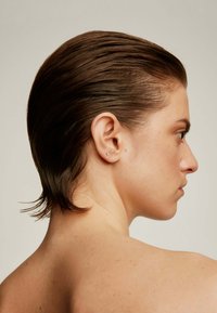 Vista de perfil de una persona con el cabello castaño oscuro peinado hacia atrás, mostrando una textura suave y mechones barridos hacia un lado. Se puede ver un pequeño pendiente de stud.