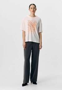 Camiseta de algodón rosa claro con mangas cortas, que presenta un gráfico floral naranja. Combinada con pantalones de pierna ancha gris oscuro y zapatos negros de punta.