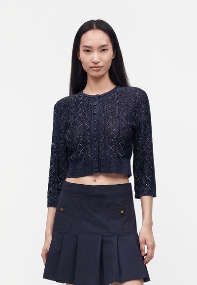 Femme portant un cardigan court bleu marine avec motif texturé et boutons, associé à une jupe plissée bleu marine avec poches avant.