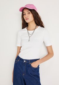 Λευκό κοντομάνικο T-shirt συνδυασμένο με σκούρο μπλε τζιν παντελόνι με ψηλή μέση και ροζ μπλεϊσμπολ καπέλο. Διαθέτει ένα στρωμένο ασημένιο κολιέ αλυσίδα.