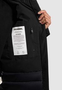Sort jakke med en glat ydre overflade, fyldt inderside, en sidelomme med lynlås og et plejeetiket med branding indeni.