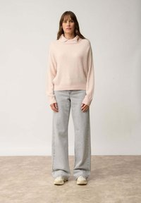Jasnoróżowy sweter z wysokim kołnierzem, długimi rękawami; zestawiony z luźnymi szarymi dżinsami i beżowymi trampkami. Miękka faktura i luźny krój.