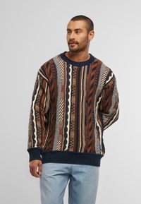 Uomo con capelli corti e barba, che indossa un maglione lavorato a maglia con motivo marrone, blu navy e crema, e jeans azzurri chiari, che guarda di lato.