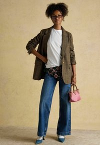 Hnedý kockovaný blazer, biele tričko, opasok so vzorom, široké modré rifle, ružová kabelka a modré lodičky. Viditeľné kučeravé vlasy a okuliare.