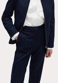 Marineblauw gestreept pak van gemêleerde stof, met een ritsjack en op maat gemaakte broek. Witte blouse met knoopsluiting eronder.