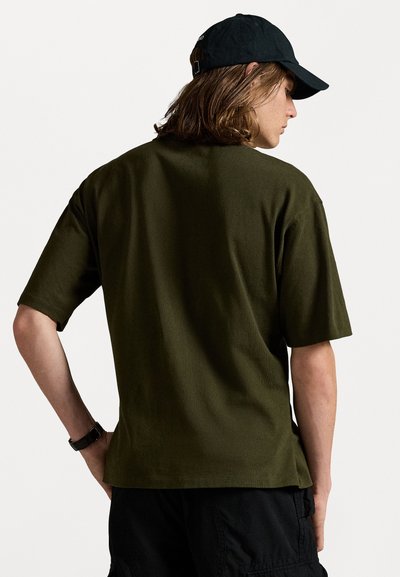Olivgrünes T-Shirt mit kurzen Ärmeln, lockerem Schnitt, weicher Textur und Rundhalsausschnitt. Kombiniert mit schwarzen Cargo-Shorts und einer schwarzen Mütze.