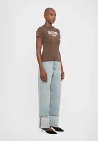 Brązowa dopasowana koszulka z napisem "MOSCHINO JEANS", zestawiona z jasnoniebieskimi szerokimi spodniami z kontrastowym przeszyciem oraz czarnymi szpilkami.
