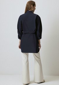 Abito camicia blu navy con colletto alto, maniche lunghe e vita aderente. Abbinato a pantaloni flare color crema e calzature chiare.