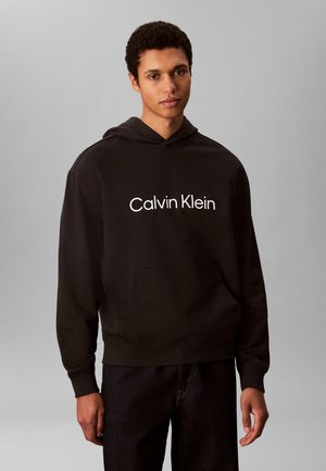 Ung mann iført en svart Calvin Klein hettegenser med frontlomme og mørke bukser, stående mot en lys grå bakgrunn.