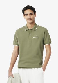 Polo shirt verde oliva in un materiale morbido, con colletto bianco e dettagli delle maniche, e una piccola tasca sul petto con dettaglio del logo.