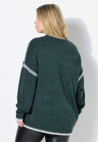 Donna con capelli biondi che indossa un maglione texturizzato verde scuro con bordi bianchi, di spalle, abbinato a pantaloni di pelle nera.