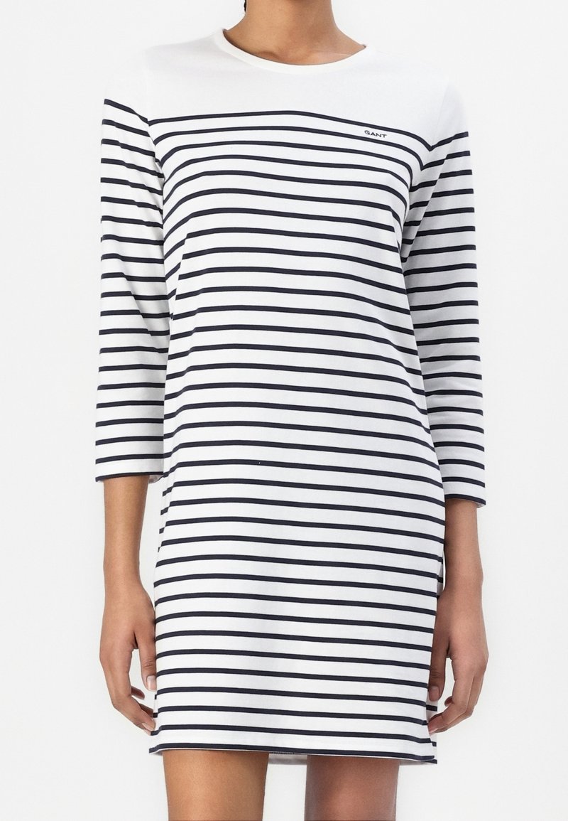 Femme portant une robe blanche avec des rayures horizontales bleu marine et des manches trois-quarts, visible du cou à la mi-cuisse.