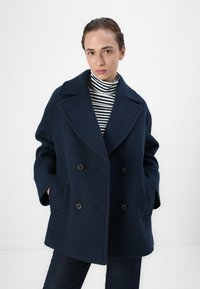 Cappotto in lana doppiopetto di colore navy con ampie rever, due tasche frontali e bottoni neri. Indossato sopra un dolcevita a righe bianche e nere.