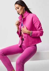 Fuchsia jack met hoge kraag en enkele knoop, gecombineerd met bijpassende fuchsia broek. Bevat een top met bloemenpatroon en zilveren sieraden.